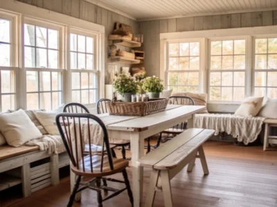 Ein gemütlicher Raum im American Cottage Style mit hell gestrichenen Holzwänden und großen Fenstern, die viel Tageslicht hereinlassen. Ein weiß gestrichener Holztisch in der Mitte des Raumes ist mit Holzbänken und Stühlen umgeben, geschmückt mit Kissen und einem Korb mit Pflanzen. An den Wänden befinden sich Regale und Dekorationsgegenstände, die zum rustikalen Charme des Stils beitragen.