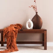 Ein Bild, das Elemente des Shaker Stils zeigt: Eine hölzerne Bank mit einem gefalteten, orangefarbenen Überwurf, eine große braune Vase mit getrockneten orangefarbenen Zweigen und eine kleine weiße Vase mit einer ovalen Öffnung. Die Gegenstände stehen vor einer hellen, einfarbigen Wand.