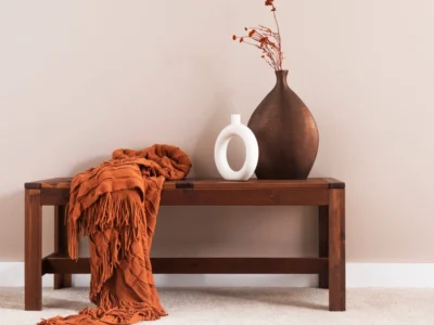 Ein Bild, das Elemente des Shaker Stils zeigt: Eine hölzerne Bank mit einem gefalteten, orangefarbenen Überwurf, eine große braune Vase mit getrockneten orangefarbenen Zweigen und eine kleine weiße Vase mit einer ovalen Öffnung. Die Gegenstände stehen vor einer hellen, einfarbigen Wand.