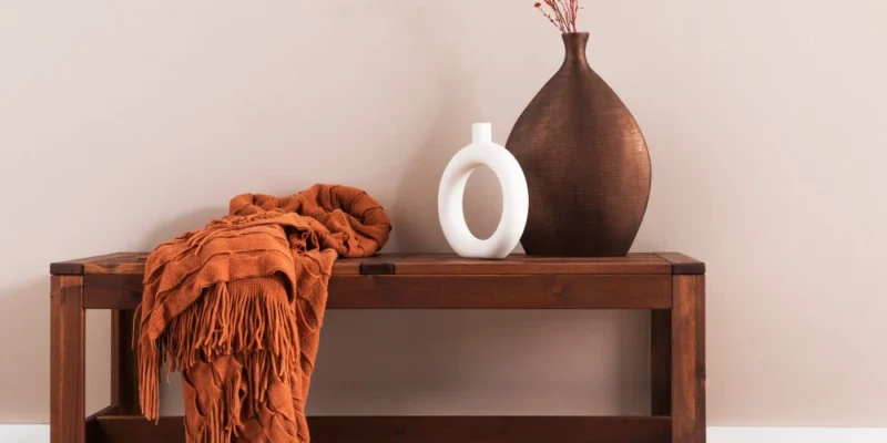 Ein Bild, das Elemente des Shaker Stils zeigt: Eine hölzerne Bank mit einem gefalteten, orangefarbenen Überwurf, eine große braune Vase mit getrockneten orangefarbenen Zweigen und eine kleine weiße Vase mit einer ovalen Öffnung. Die Gegenstände stehen vor einer hellen, einfarbigen Wand.