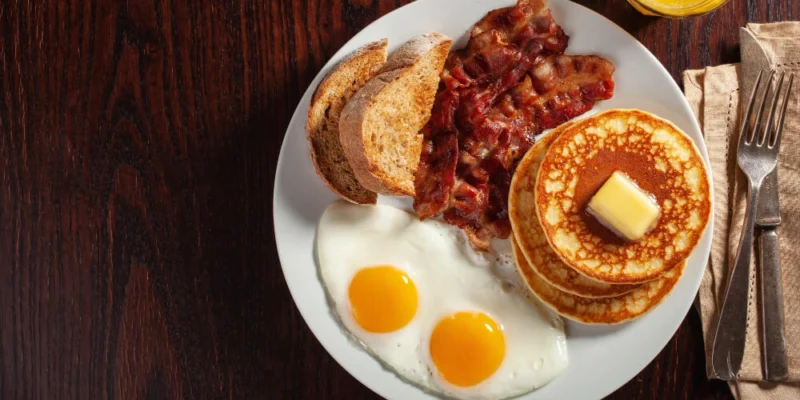 Ein klassisches amerikanisches Frühstück auf einem Teller, bestehend aus zwei Spiegeleiern mit flüssigem Dotter, knusprigem Speck, zwei Scheiben getoastetem Brot und einem Stapel goldbrauner Pancakes mit einem Stück Butter und Ahornsirup. Ein Glas Orangensaft und Besteck vervollständigen die Mahlzeit, die auf einem dunklen Holztisch serviert wird.