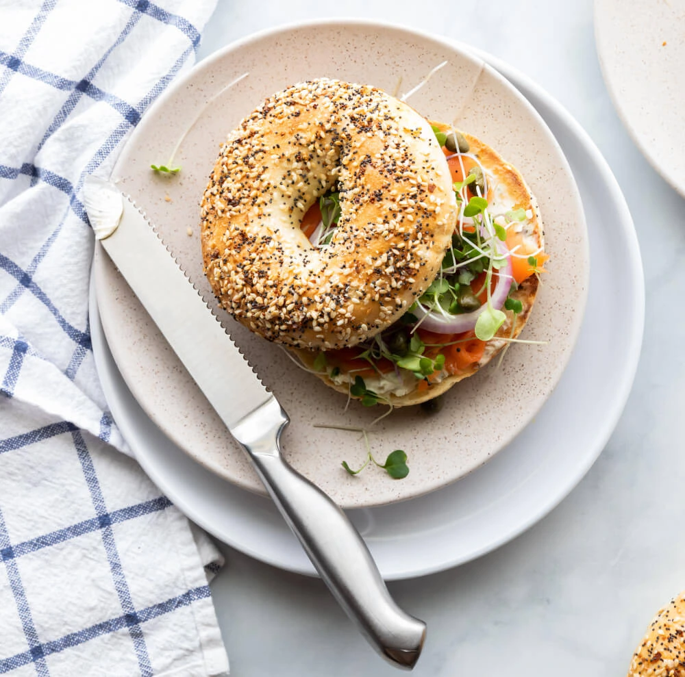 Ein aufgeschnittener "Everything Bagel" mit Lachs, Frischkäse, roten Zwiebeln, Kapern und frischen Microgreens, eine Variante des amerikanischen Frühstücks, wird aus der Vogelperspektive auf einem cremefarbenen Teller gezeigt. Ein Messer mit silberner Klinge und metallenem Griff liegt diagonal über dem Teller. Ein blau-weiß kariertes Tuch mit sichtbarer Textur ist links vom Teller zu sehen.