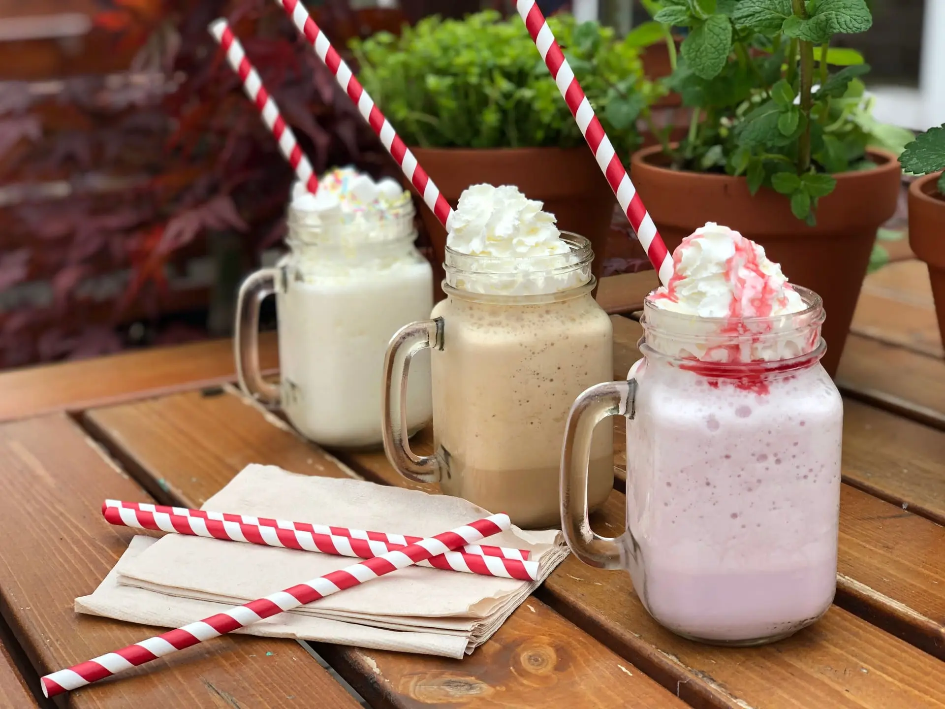 Drei leckere Milchshakes in Mason-Gläsern stehen auf einem rustikalen Holztisch im Freien. Jeder Shake hat eine andere Farbe – Vanille, Kaffee und Erdbeere – und ist mit einer großzügigen Portion Schlagsahne garniert. Der vordere Erdbeer-Shake hat zusätzlich eine rote Sirup-Dekoration. In jedem Glas steckt ein rot-weiß gestreifter Papierstrohhalm. Im Hintergrund stehen grüne Kräutertöpfe, die eine frische und gemütliche Atmosphäre schaffen. Auf dem Tisch liegen zudem Servietten und zwei zusätzliche Strohhalme.