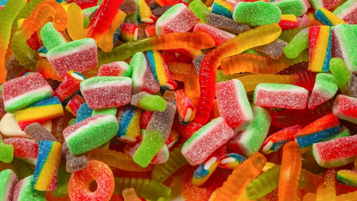 US-Süßigkeiten in verschiedenen Formen, darunter saure Wassermelonenstücke, Regenbogen-Streifen und Gummi-Würmer. Die Süßigkeiten sind mit Zucker bestreut und liegen dicht beieinander, was eine lebendige und farbenfrohe Szene ergibt.