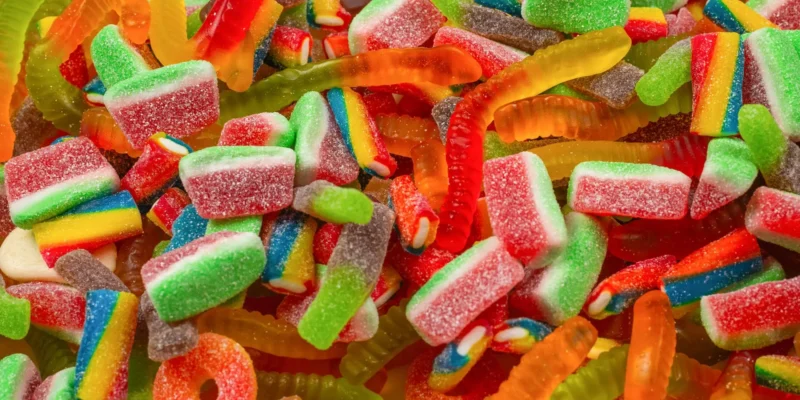 US-Süßigkeiten in verschiedenen Formen, darunter saure Wassermelonenstücke, Regenbogen-Streifen und Gummi-Würmer. Die Süßigkeiten sind mit Zucker bestreut und liegen dicht beieinander, was eine lebendige und farbenfrohe Szene ergibt.
