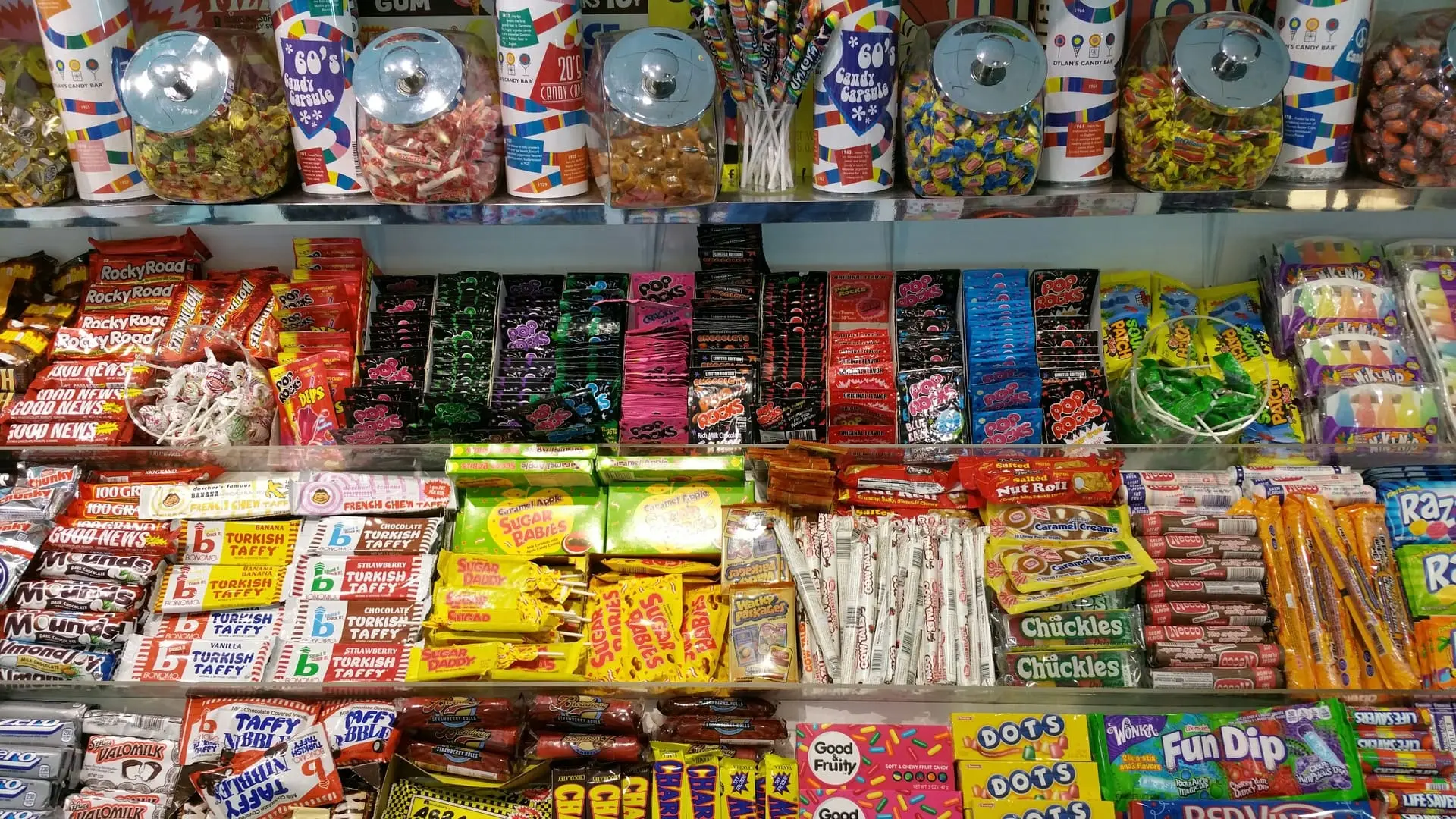 Ein farbenfrohes Süßigkeitenregal in einem Laden, gefüllt mit einer Vielzahl von amerikanischen Süßigkeiten, darunter Schokoriegel, Kaubonbons, Lutscher und Knisterbonbons. Gläser mit Bonbons stehen auf der oberen Ablage, während in den unteren Reihen bekannte Marken wie Pop Rocks, Turkish Taffy und Good News zu sehen sind. Die Szene vermittelt ein Gefühl von Nostalgie und Süßigkeitenvielfalt. Weniger anzeigen
