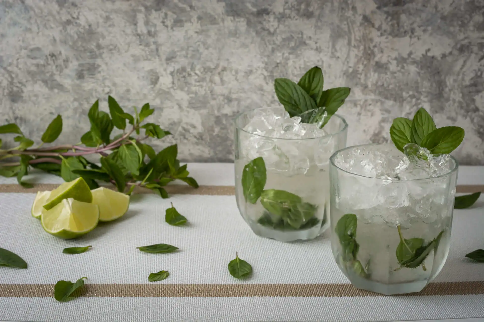 Zwei erfrischende Mint Julep-Cocktails in kristallklaren Gläsern, serviert mit frischer Minze, Limettenscheiben und viel Crushed Ice. Auf dem Tisch liegen Minzzweige und Limetten, was dem Bild eine natürliche und sommerliche Atmosphäre verleiht.