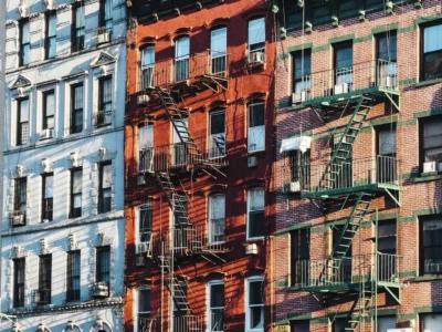 Fassaden mehrerer Altbauten mit Feuerleitern in einer städtischen Umgebung. Die Gebäude haben unterschiedliche Farben, darunter Blau, Rot und Backstein mit grünen Fensterrahmen. Die Metall-Feuertreppen werfen lange Schatten auf die Fassaden, was die typische Architektur vieler New Yorker Wohnhäuser widerspiegelt.