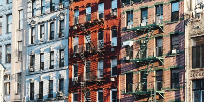 Fassaden mehrerer Altbauten mit Feuerleitern in einer städtischen Umgebung. Die Gebäude haben unterschiedliche Farben, darunter Blau, Rot und Backstein mit grünen Fensterrahmen. Die Metall-Feuertreppen werfen lange Schatten auf die Fassaden, was die typische Architektur vieler New Yorker Wohnhäuser widerspiegelt.