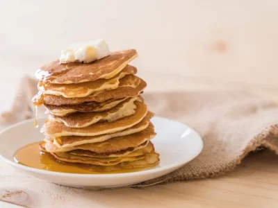 Ein hoher Stapel goldbrauner Pfannkuchen auf einem weißen Teller, gekrönt von einem Stück Butter. Ahornsirup fließt langsam über die Pfannkuchen und sammelt sich auf dem Teller. Im Hintergrund liegt ein grob gewebtes Leinentuch auf einer hellen Holzoberfläche.