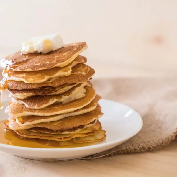Ein hoher Stapel goldbrauner Pfannkuchen auf einem weißen Teller, gekrönt von einem Stück Butter. Ahornsirup fließt langsam über die Pfannkuchen und sammelt sich auf dem Teller. Im Hintergrund liegt ein grob gewebtes Leinentuch auf einer hellen Holzoberfläche.