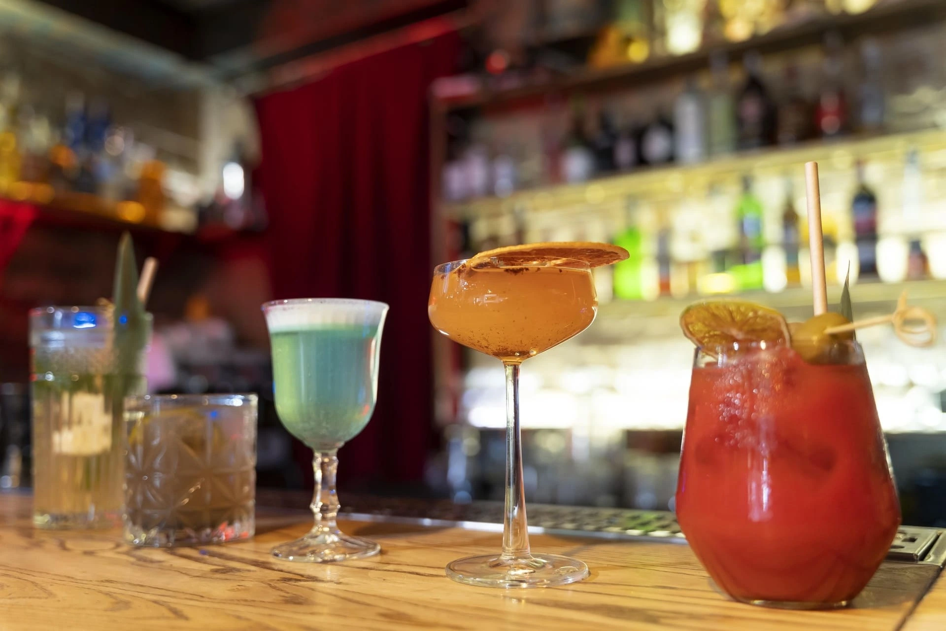 Eine stilvolle Bar-Szene mit fünf typischen Amerikanischen Cocktails, die auf einer hölzernen Theke stehen. Jedes Getränk ist in einem einzigartigen Glas serviert und mit kreativen Garnituren verziert, darunter getrocknete Früchte, Oliven und Kräuter. Im Hintergrund ist eine beleuchtete Bar mit Flaschen und Regalen sichtbar, während rote Vorhänge eine gemütliche Atmosphäre schaffen. Die Szene vermittelt einen eleganten und einladenden Eindruck, perfekt für einen entspannten Abend in einer gehobenen Bar.