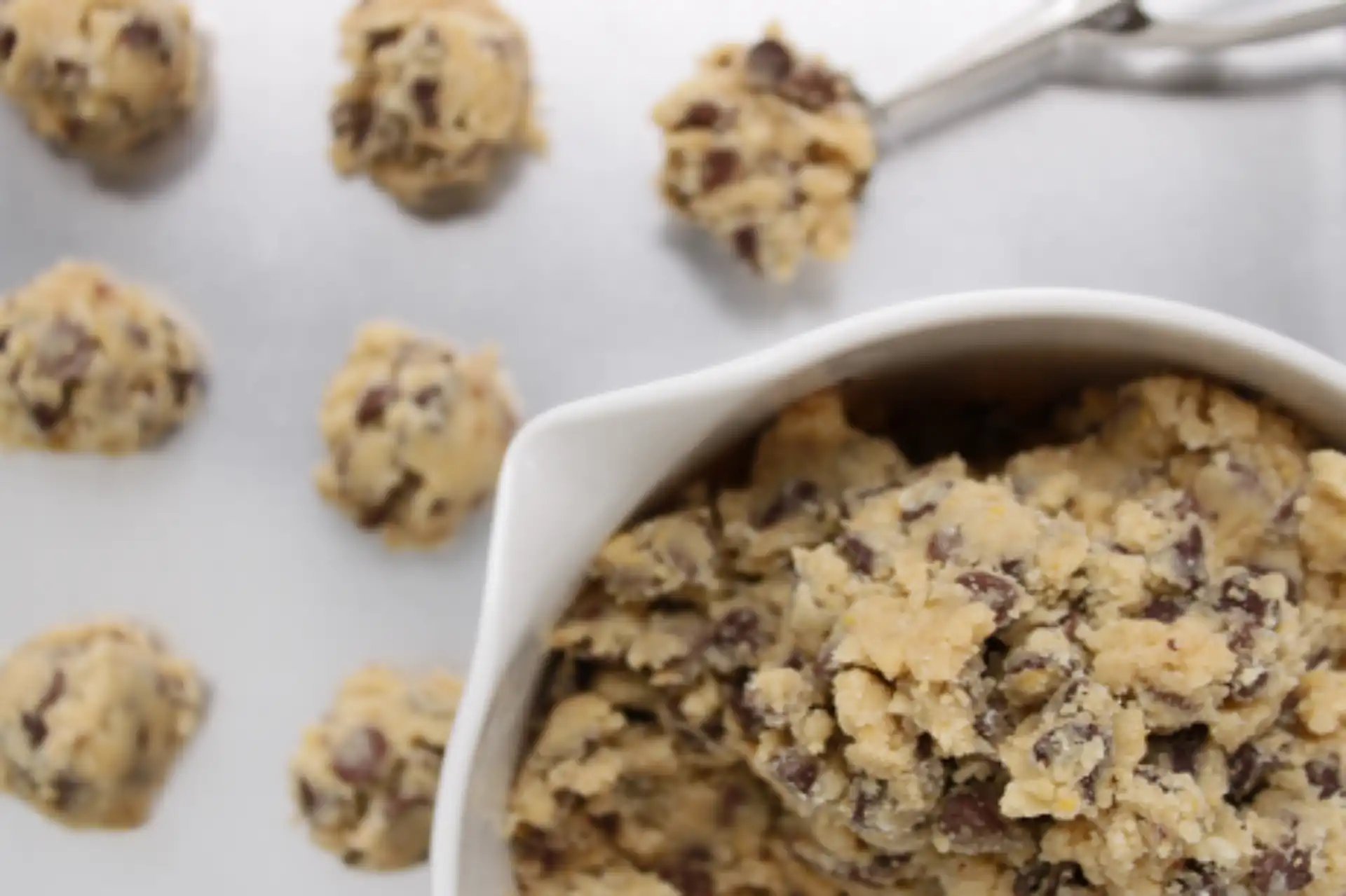 Nahaufnahme von rohem Chocolate-Chip-Cookie-Teig in einer weißen Schüssel, daneben mehrere Teigkugeln auf einem Tisch. Im Hintergrund ist ein Portionierer mit weiterer Teigkugel zu sehen – bereit für den Ofen.