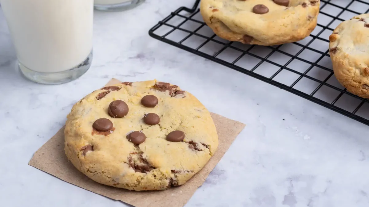 Frische, dicke american Chocolate-Chip-Cookies auf einem Marmortisch, begleitet von einem Glas Milch. Ein Cookie liegt auf braunem Papier im Vordergrund, weitere Cookies kühlen auf einem schwarzen Gitterrost im Hintergrund.