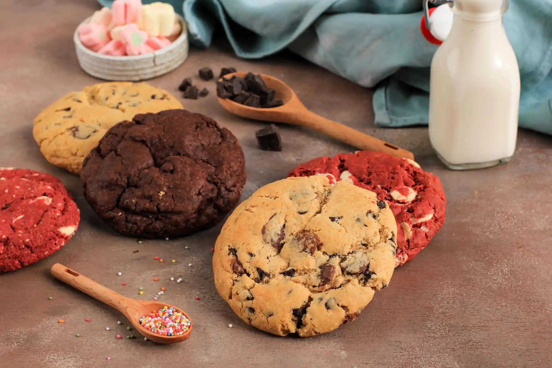 Verschiedene Sorten großer, weicher Cookies – darunter Schoko-, Red-Velvet- und Chocolate-Chip-Cookies – liegen auf einem rustikalen Holztisch. Im Hintergrund befinden sich eine Milchflasche, Holzlöffel mit Schokostückchen und Zuckerstreuseln sowie eine Schale mit Marshmallows.
