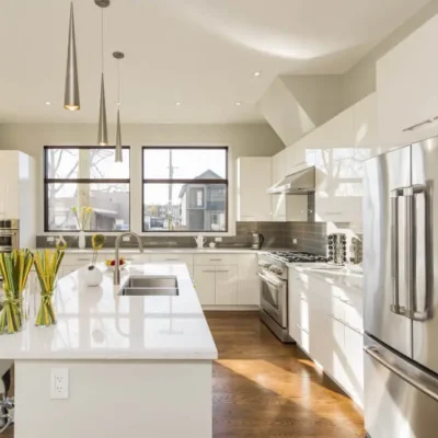 Eine helle, geräumige offene Küche im amerikanischen Stil mit einer zentralen Kücheninsel und modernen weißen Schränken. Die Küche ist gut beleuchtet mit großen Fenstern und Hängelampen über der Insel. Edelstahlgeräte, einschließlich eines großen Kühlschranks, ergänzen das saubere, moderne Design.