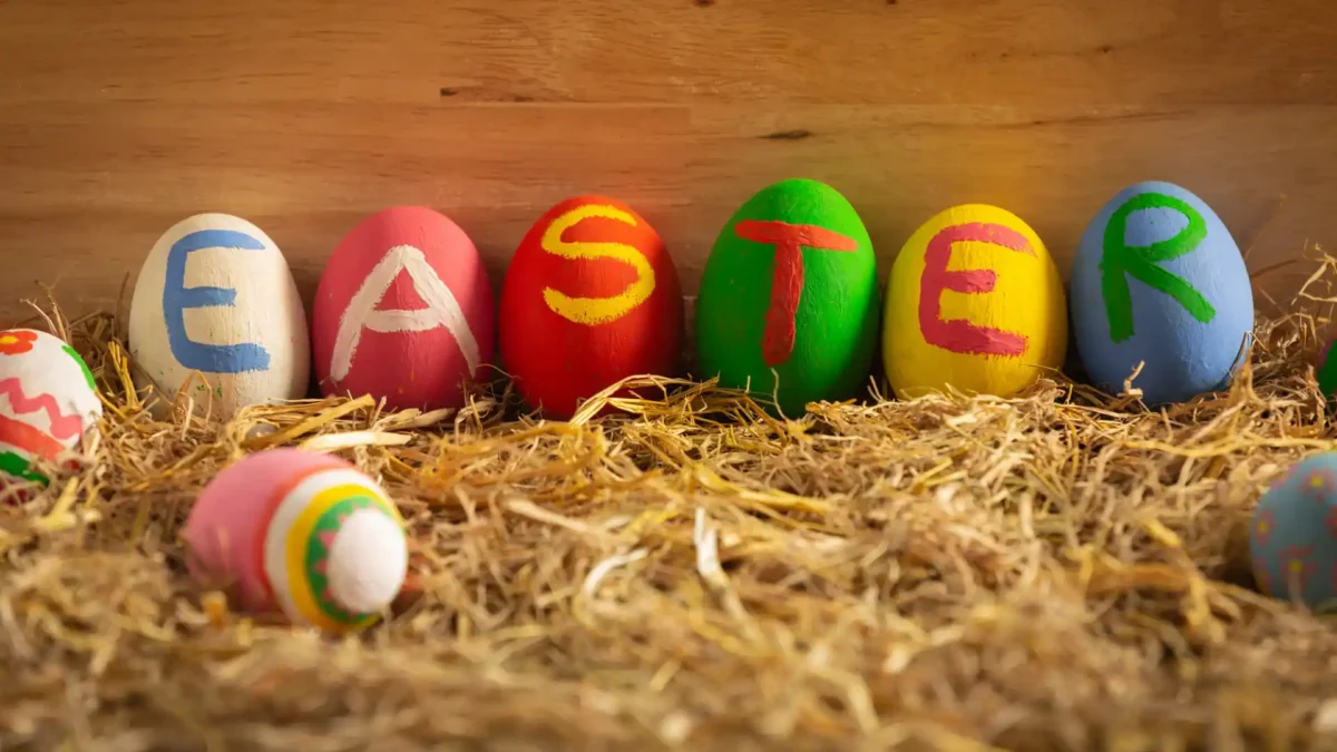 Bunte bemalte Ostereier mit dem Schriftzug ‚EASTER‘ auf Stroh vor Holz-Hintergrund – kreative Osterdeko.