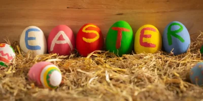 Bunte bemalte Ostereier mit dem Schriftzug ‚EASTER‘ auf Stroh vor Holz-Hintergrund – kreative Osterdeko.