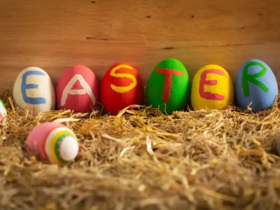 Bunte bemalte Ostereier mit dem Schriftzug ‚EASTER‘ auf Stroh vor Holz-Hintergrund – kreative Osterdeko.