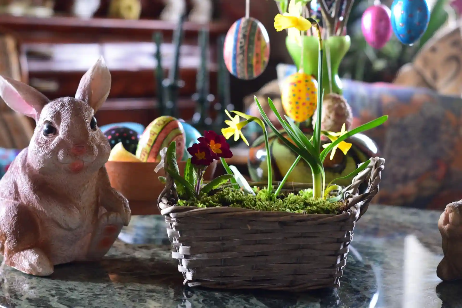 Osterdekoration mit Keramik-Osterhase, bunten Ostereiern und Frühlingsblumen in einem geflochtenen Korb auf einem Tisch.