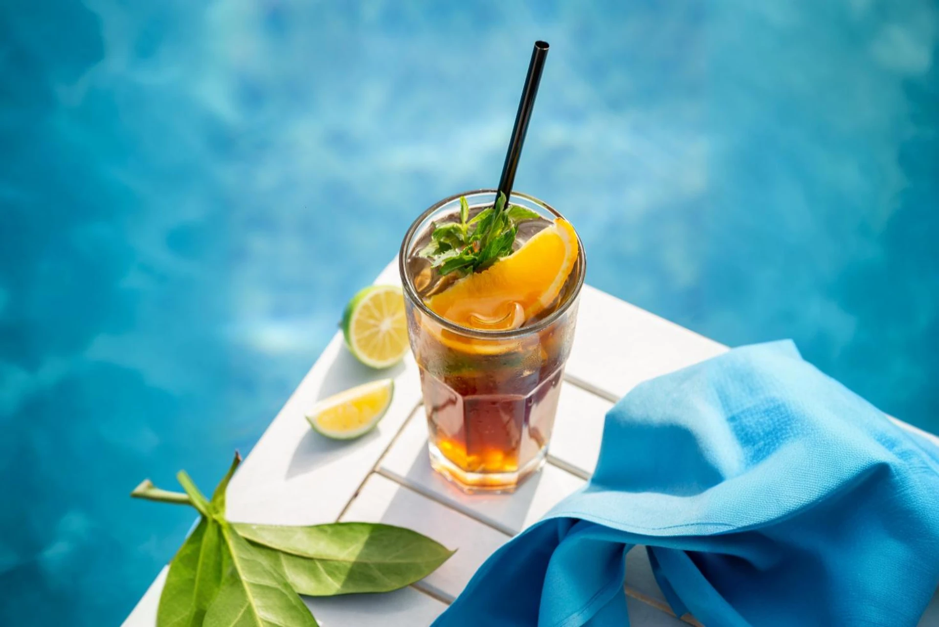 Ein belebender Arnold Palmer, eine Mischung aus Eistee und Limonade, wird in einem hohen Glas mit Eiswürfeln, einer Orangenspalte und Minze präsentiert. Ein schwarzer Strohhalm ragt aus dem Getränk. Das Glas steht auf einer weißen, lattenartigen Oberfläche neben einem gefalteten, hellblauen Tuch und einigen grünen Blättern. Im Hintergrund verschwimmt das leuchtend blaue Wasser eines Pools, was die erfrischende und sommerliche Assoziation des Arnold Palmer verstärkt.