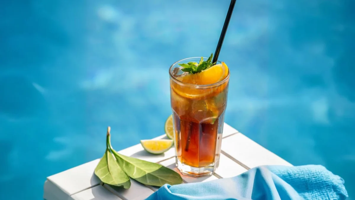 Ein erfrischender Arnold Palmer, erkennbar an seiner hellbraunen Farbe und den Zitrusfruchtscheiben, steht in einem hohen Glas mit einem schwarzen Strohhalm. Das Glas ist mit Minze und Orangenspalten garniert und befindet sich auf einer weißen Oberfläche, die an einen Liegestuhl erinnert. Im Hintergrund ist das verschwommene, azurblaue Wasser eines Pools zu sehen, was die sommerliche und entspannte Atmosphäre des Getränks unterstreicht.