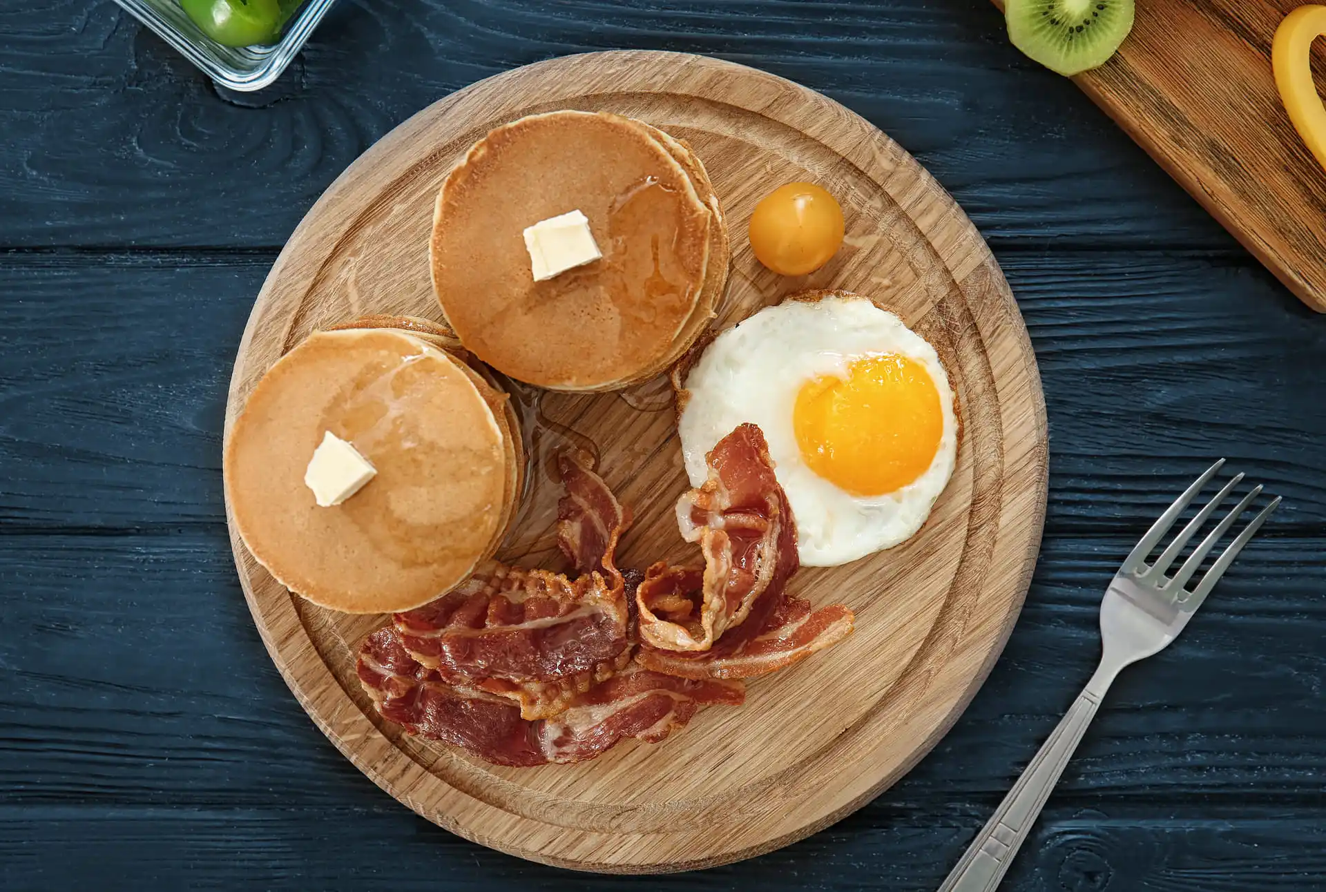 Frühstücksteller mit zwei gestapelten Pancakes, jeweils mit einem Stück Butter und Ahornsirup, dazu knuspriger Bacon, ein Spiegelei und eine gelbe Kirschtomate auf einem runden Holzbrett, angerichtet auf dunklem Holztisch.