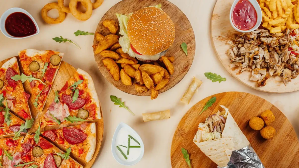 Ein appetitliches Flat-Lay präsentiert eine vielfältige Auswahl an Fast Food Gerichten, darunter Burger, Döner, Pizza und Zwiebelringe. Die Speisen sind ansprechend auf einem hellen Untergrund arrangiert und laden zum Zugreifen ein.