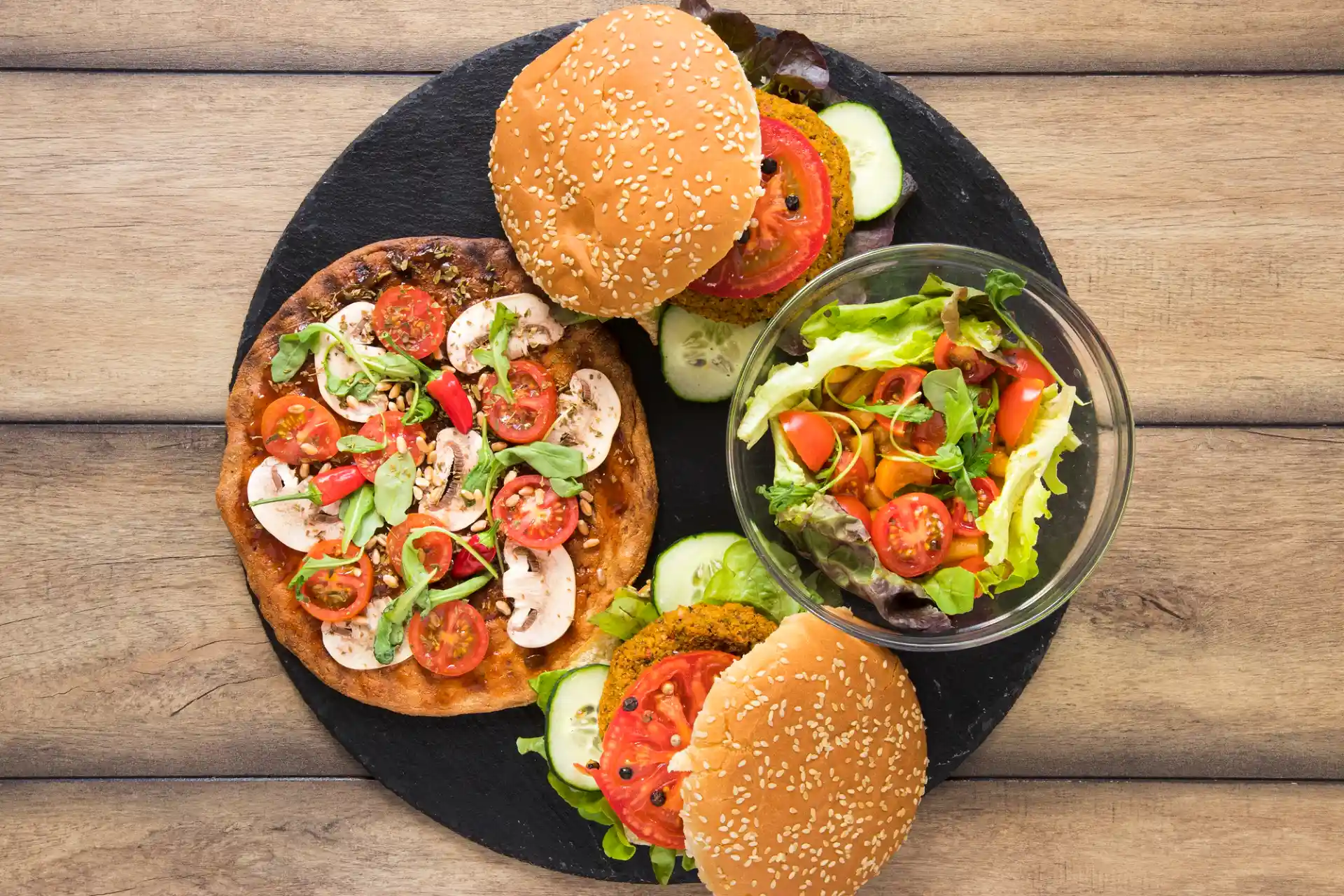 Ein Flat-Lay zeigt eine Auswahl an Fast Food in einer gesünderen Variante: zwei Veggie Burger mit Salat und Tomaten, eine kleine Pizza mit Gemüse und Kräutern sowie eine Schüssel frischen Salat. Die Speisen sind auf einem dunklen Teller platziert, der auf einem hölzernen Untergrund liegt.