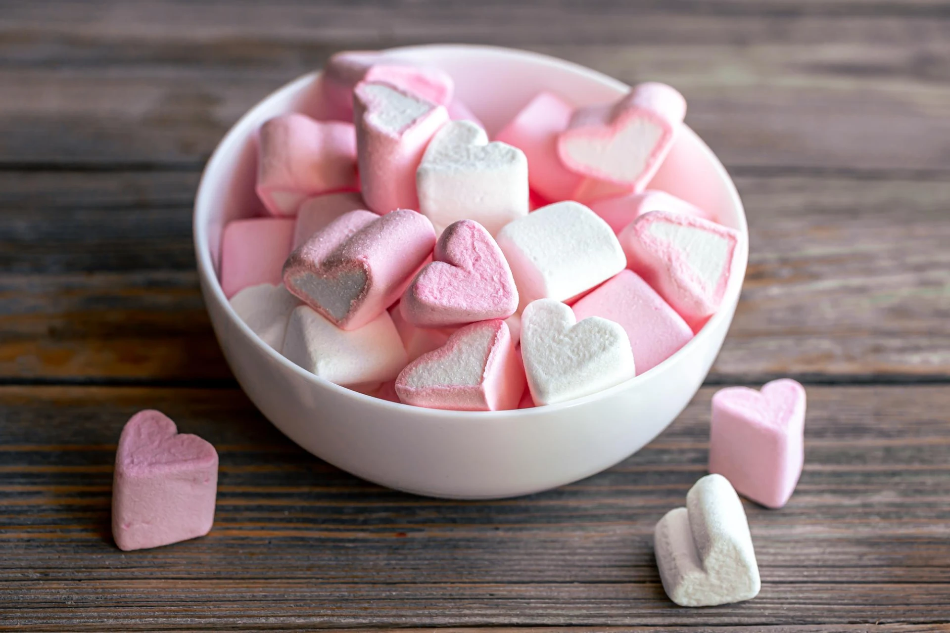 Ein weißes Schälchen ist prall gefüllt mit rosa und weißen herzförmigen Marshmallows. Einige einzelne Marshmallows in Herzform liegen verstreut auf einem rustikalen Holztisch. Die süßen Leckereien wirken einladend und könnten auf eine Feier oder einen besonderen Anlass hindeuten.