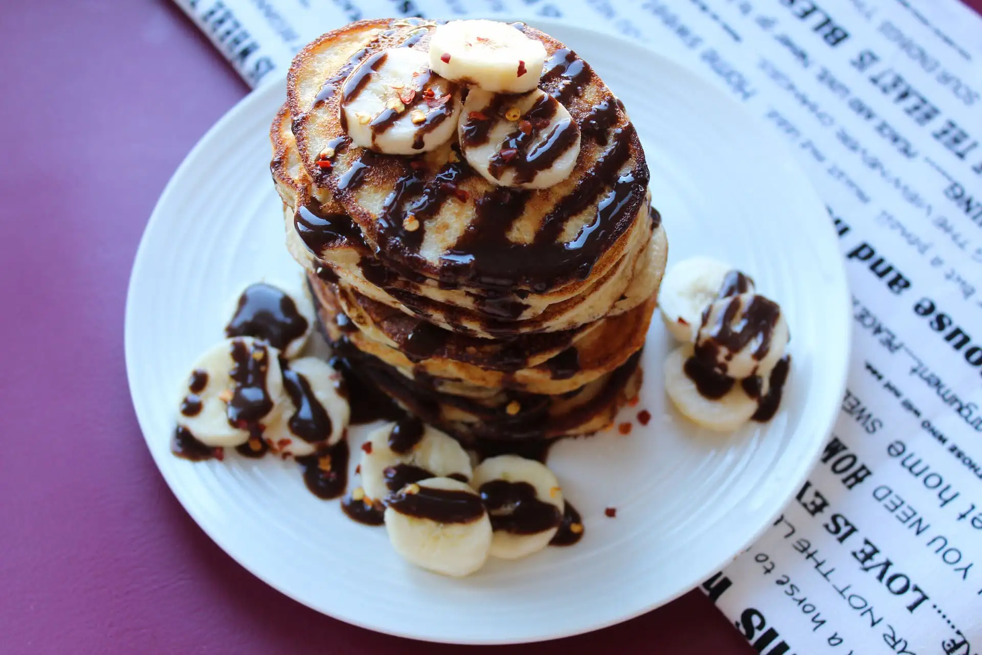 Auf dem Bild ist ein Teller mit einem Stapel Pancakes zu sehen. Die Pancakes sind mit Bananenscheiben, Schokoladensoße und einer Prise Chiliflocken garniert. Um den Stapel herum liegen weitere Bananenscheiben, ebenfalls mit Schokosoße überzogen. Der Teller steht auf einer Oberfläche in Dunkelrot, daneben liegt eine Serviette mit schwarzem Textdruck.