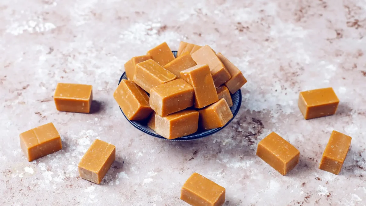 Eine kleine, dunkle Schale ist gefüllt mit rechteckigen, hellbraunen Karamellbonbons. Weitere einzelne Karamellbonbons sind um die Schale herum auf einer hellbraun-grau gesprenkelten Oberfläche verstreut. Die Draufsicht betont die weiche Textur und die einheitliche Farbe der süßen Leckereien.