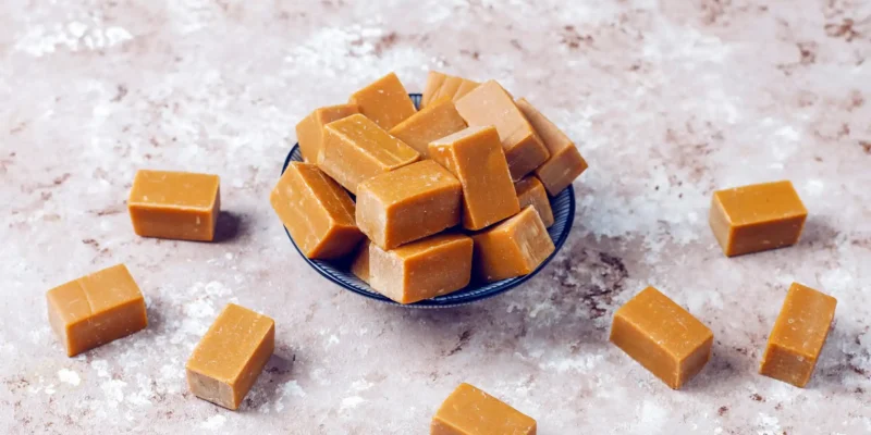 Eine kleine, dunkle Schale ist gefüllt mit rechteckigen, hellbraunen Karamellbonbons. Weitere einzelne Karamellbonbons sind um die Schale herum auf einer hellbraun-grau gesprenkelten Oberfläche verstreut. Die Draufsicht betont die weiche Textur und die einheitliche Farbe der süßen Leckereien.