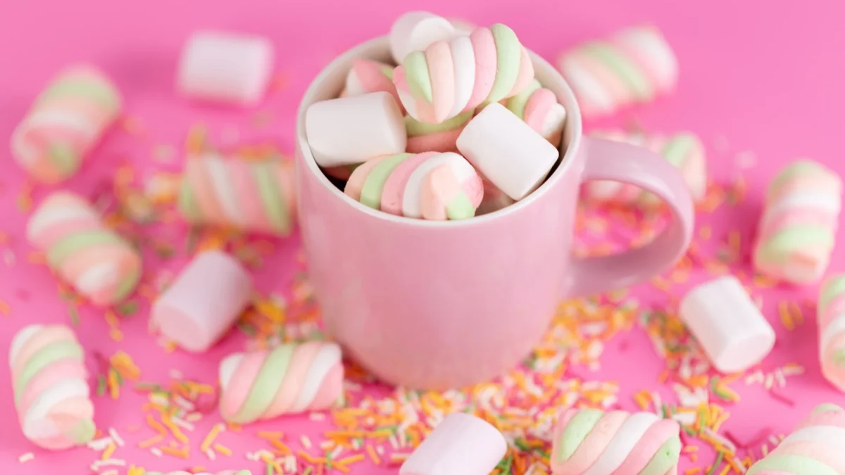 Eine hellrosa Tasse ist bis zum Rand gefüllt mit weißen und spiralförmig rosa-grün gefärbten Marshmallows. Um die Tasse herum sind weitere Marshmallows und bunte Zuckerstreusel auf einem leuchtend pinken Hintergrund verstreut. Die süße und farbenfrohe Szene wirkt festlich und einladend.