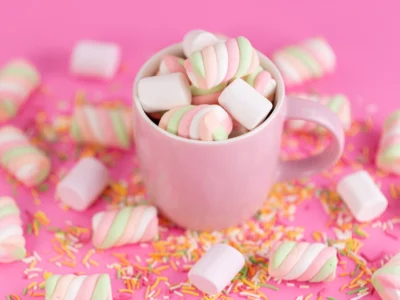 Eine hellrosa Tasse ist bis zum Rand gefüllt mit weißen und spiralförmig rosa-grün gefärbten Marshmallows. Um die Tasse herum sind weitere Marshmallows und bunte Zuckerstreusel auf einem leuchtend pinken Hintergrund verstreut. Die süße und farbenfrohe Szene wirkt festlich und einladend.