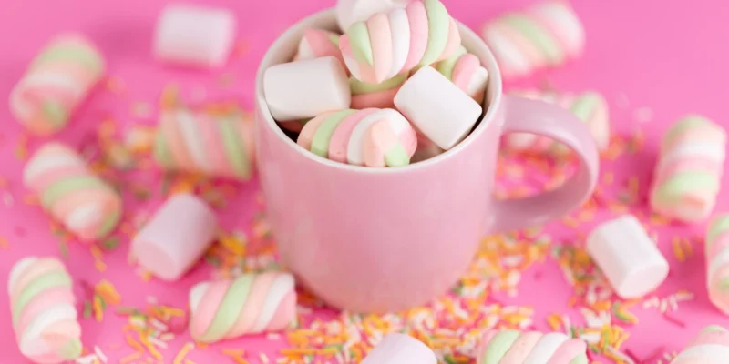 Eine hellrosa Tasse ist bis zum Rand gefüllt mit weißen und spiralförmig rosa-grün gefärbten Marshmallows. Um die Tasse herum sind weitere Marshmallows und bunte Zuckerstreusel auf einem leuchtend pinken Hintergrund verstreut. Die süße und farbenfrohe Szene wirkt festlich und einladend.