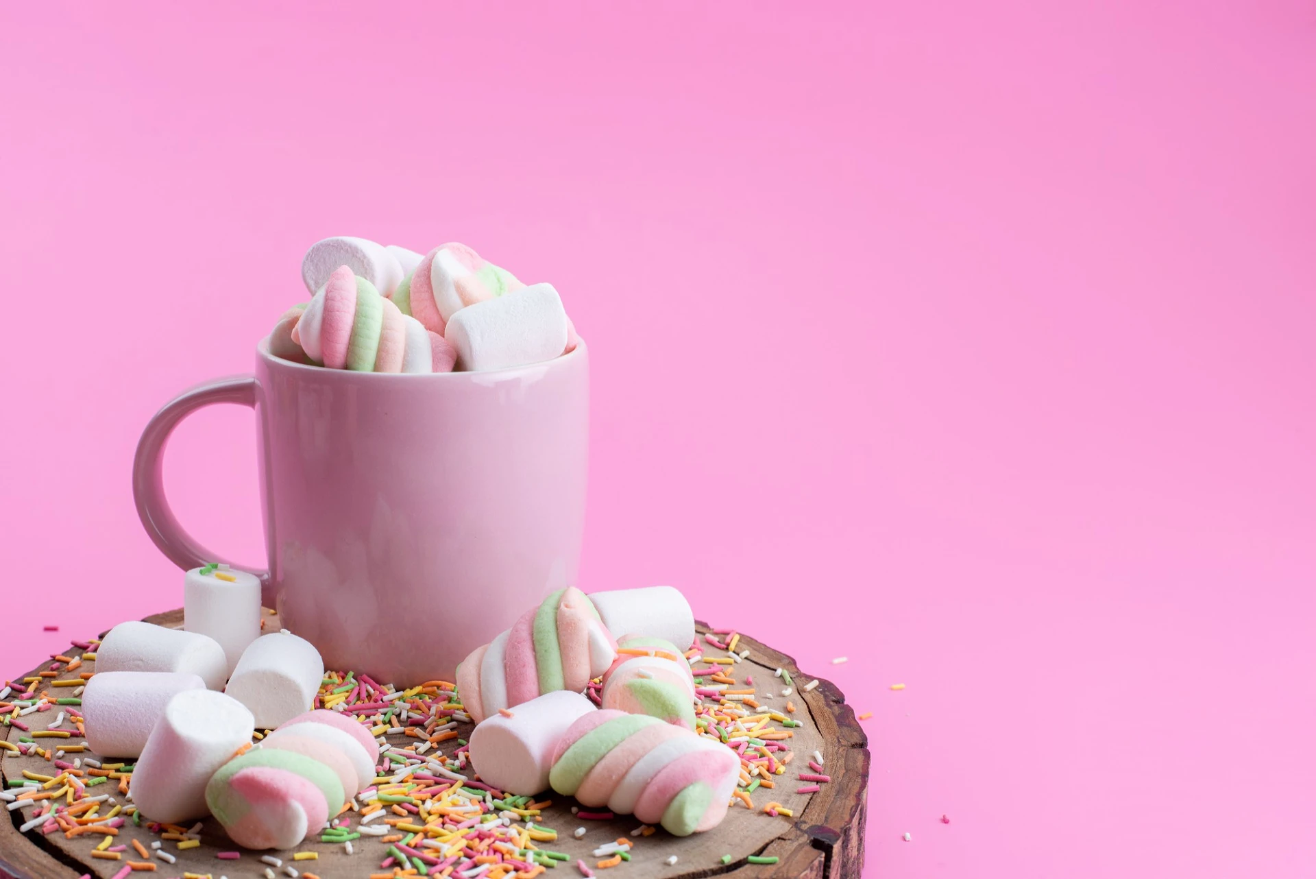 Eine hellrosa Tasse, gefüllt mit weißen und spiralförmig rosa-grün gefärbten Marshmallows, steht auf einer rustikalen Holzscheibe. Weitere Marshmallows und bunte Zuckerstreusel sind um die Tasse herum auf der Holzscheibe und dem leuchtend pinken Hintergrund verstreut. Die süße und farbenfrohe Komposition wirkt verspielt und einladend.