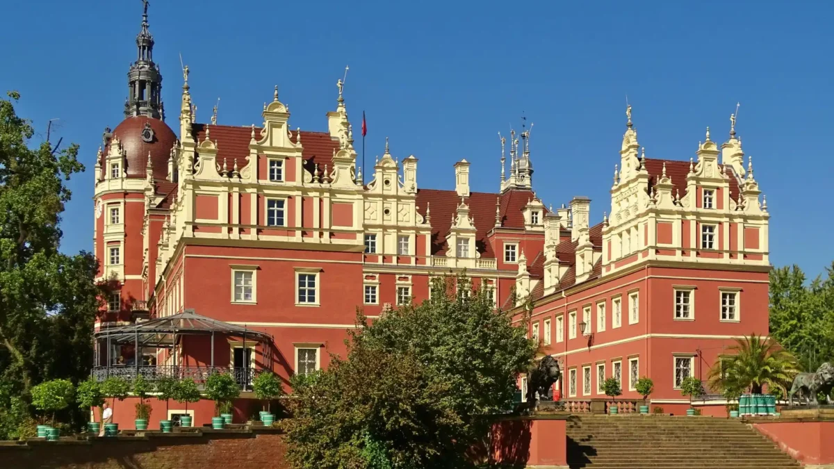 Rückansicht des Schlosses im Fürst-Pückler-Park Bad Muskau bei strahlend blauem Himmel – beeindruckende Architektur mit barocken Elementen, roten Fassaden, Türmchen und verzierten Giebeln, flankiert von Löwenstatuen und gepflegter Gartenanlage.