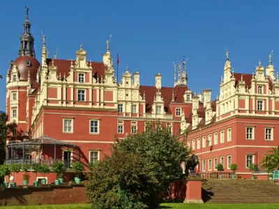 Rückansicht des Schlosses im Fürst-Pückler-Park Bad Muskau bei strahlend blauem Himmel – beeindruckende Architektur mit barocken Elementen, roten Fassaden, Türmchen und verzierten Giebeln, flankiert von Löwenstatuen und gepflegter Gartenanlage.