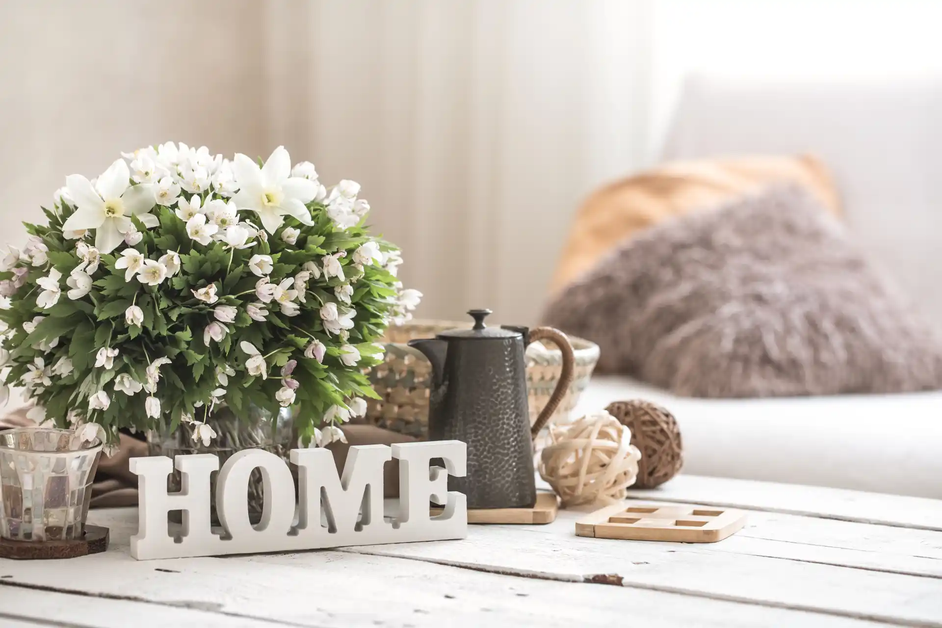Stillleben im Wohnzimmer im Country Style: Rustikaler Holztisch mit liebevoll arrangiertem Blumenstrauß, antiker Kaffeekanne und handgeschriebenem Schriftzug ‚HOME‘.