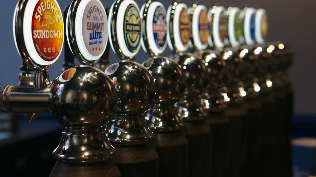 Nahaufnahme einer glänzenden Reihe von Bierzapfhähnen mit beleuchteten Logos verschiedener Biersorten, darunter Speight’s Sundown und Summit Ultra. Die Zapfhähne sind aus poliertem Metall und bilden eine gleichmäßige Linie auf einer Theke in einer Bar oder Brauerei.