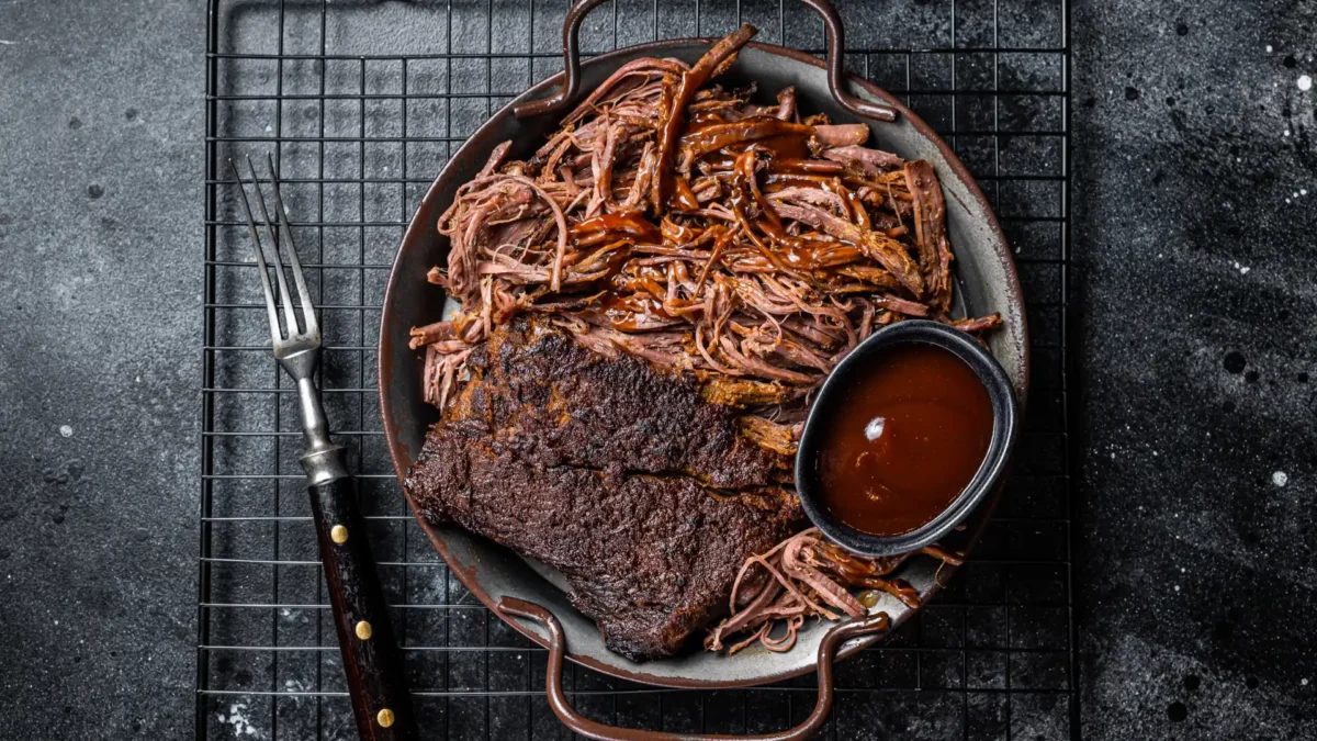 Draufsicht auf einen Teller mit zartem, langsam gegartem Pulled Beef, teilweise fein zerzupft und mit Barbecue-Soße übergossen. Daneben liegt ein Stück geräuchertes Brisket mit würziger Kruste. Eine kleine Schale mit BBQ-Soße und eine Fleischgabel runden das rustikale Arrangement auf einem dunklen Gitterrost ab.