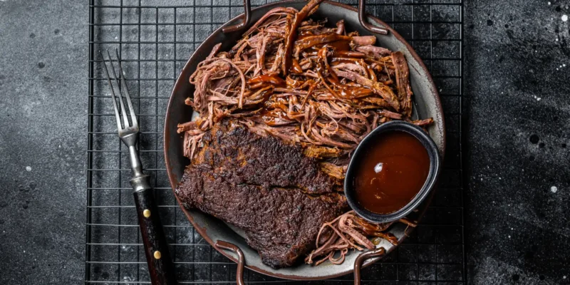 Draufsicht auf einen Teller mit zartem, langsam gegartem Pulled Beef, teilweise fein zerzupft und mit Barbecue-Soße übergossen. Daneben liegt ein Stück geräuchertes Brisket mit würziger Kruste. Eine kleine Schale mit BBQ-Soße und eine Fleischgabel runden das rustikale Arrangement auf einem dunklen Gitterrost ab.