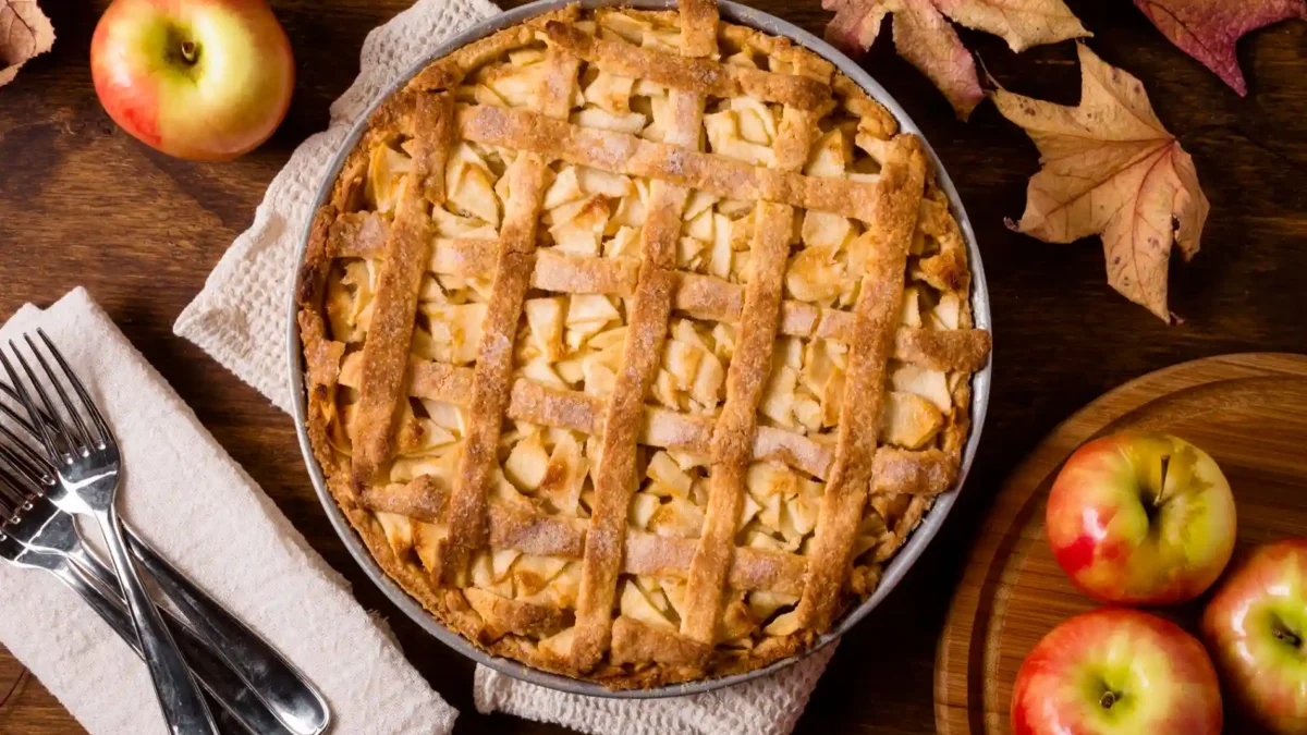 Ein rustikales von oben aufgenommenes Bild zeigt einen frisch gebackenen Apple Pie in einer runden Backform, mittig auf einem grob gewebten, hellen Küchentuch platziert. Der goldbraune Kuchen ist mit einem Gittermuster aus Teigstreifen bedeckt, unter dem Apfelstücke hervorblitzen. Rund um den Kuchen sind Herbstakzente arrangiert: Links neben dem Küchentuch liegen drei silberne Gabeln auf einem weiteren hellen Stofftuch. Oben links und unten rechts befinden sich einzelne leuchtend rote und gelbe Äpfel, einige davon auf einem runden Holzbrett. Trockene, herbstliche Ahornblätter in Rottönen sind über den oberen rechten Bildbereich verteilt und verstärken die gemütliche Herbststimmung.