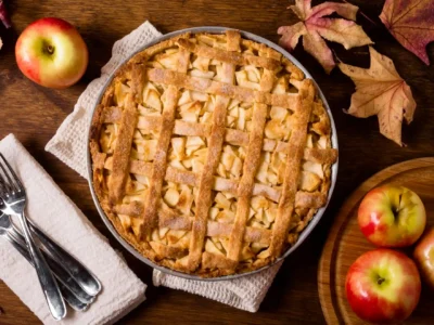 Ein rustikales von oben aufgenommenes Bild zeigt einen frisch gebackenen Apple Pie in einer runden Backform, mittig auf einem grob gewebten, hellen Küchentuch platziert. Der goldbraune Kuchen ist mit einem Gittermuster aus Teigstreifen bedeckt, unter dem Apfelstücke hervorblitzen. Rund um den Kuchen sind Herbstakzente arrangiert: Links neben dem Küchentuch liegen drei silberne Gabeln auf einem weiteren hellen Stofftuch. Oben links und unten rechts befinden sich einzelne leuchtend rote und gelbe Äpfel, einige davon auf einem runden Holzbrett. Trockene, herbstliche Ahornblätter in Rottönen sind über den oberen rechten Bildbereich verteilt und verstärken die gemütliche Herbststimmung.