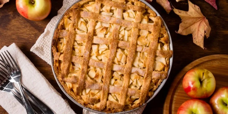 Ein rustikales von oben aufgenommenes Bild zeigt einen frisch gebackenen Apple Pie in einer runden Backform, mittig auf einem grob gewebten, hellen Küchentuch platziert. Der goldbraune Kuchen ist mit einem Gittermuster aus Teigstreifen bedeckt, unter dem Apfelstücke hervorblitzen. Rund um den Kuchen sind Herbstakzente arrangiert: Links neben dem Küchentuch liegen drei silberne Gabeln auf einem weiteren hellen Stofftuch. Oben links und unten rechts befinden sich einzelne leuchtend rote und gelbe Äpfel, einige davon auf einem runden Holzbrett. Trockene, herbstliche Ahornblätter in Rottönen sind über den oberen rechten Bildbereich verteilt und verstärken die gemütliche Herbststimmung.