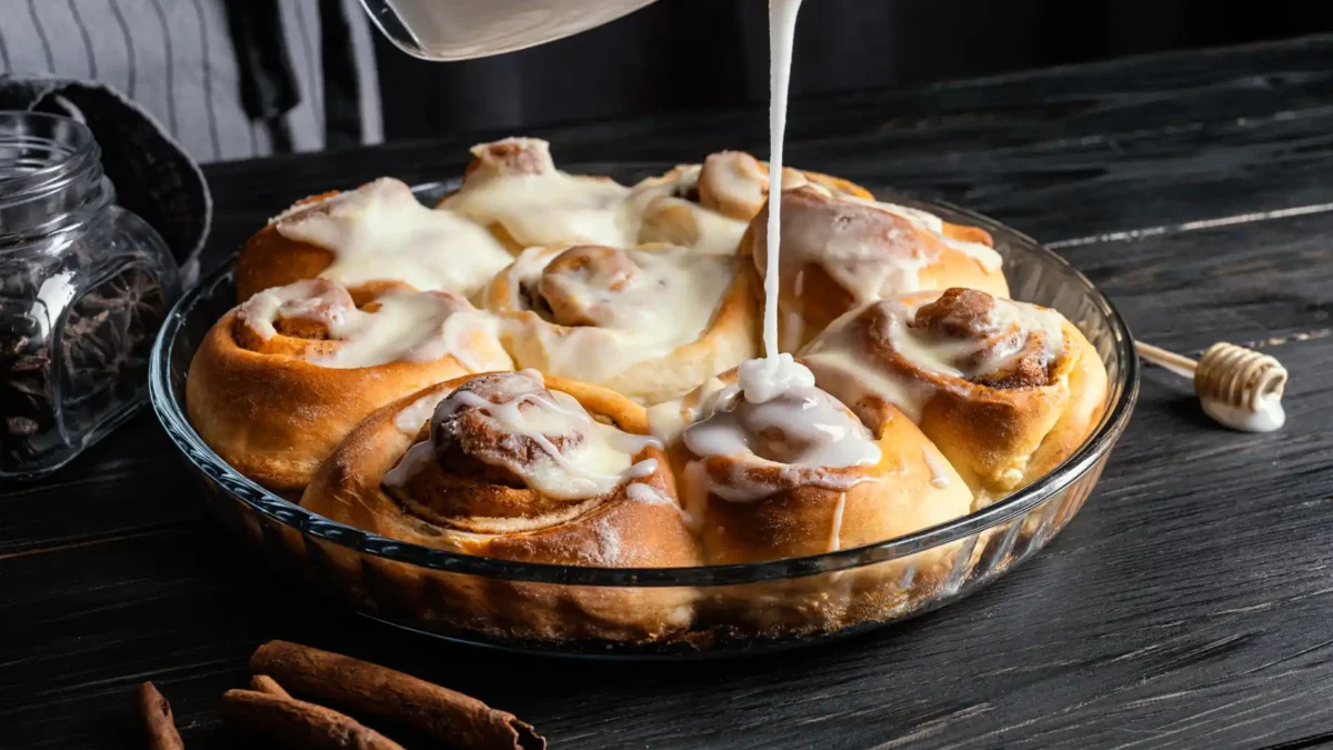 Ein hochauflösendes, dunkles Stimmungsbild zeigt eine Glasform, die mit frisch gebackenen, glasierten Cinnamon Rolls gefüllt ist. Eine Hand gießt aus einem durchsichtigen Gefäß eine dicke, weiße Glasur über die zimtigen Teigschnecken, wodurch der Guss auf einigen bereits glasierten Rolls glänzt. Auf dem rustikalen, dunklen Holztisch liegen Zimtstangen, ein Holzlöffel mit Honig und ein kleines Glasgefäß mit Kaffeebohnen, die das aromatische Thema unterstreichen.