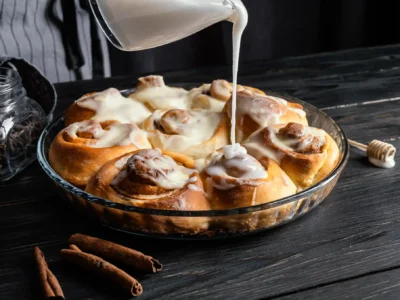 Ein hochauflösendes, dunkles Stimmungsbild zeigt eine Glasform, die mit frisch gebackenen, glasierten Cinnamon Rolls gefüllt ist. Eine Hand gießt aus einem durchsichtigen Gefäß eine dicke, weiße Glasur über die zimtigen Teigschnecken, wodurch der Guss auf einigen bereits glasierten Rolls glänzt. Auf dem rustikalen, dunklen Holztisch liegen Zimtstangen, ein Holzlöffel mit Honig und ein kleines Glasgefäß mit Kaffeebohnen, die das aromatische Thema unterstreichen.