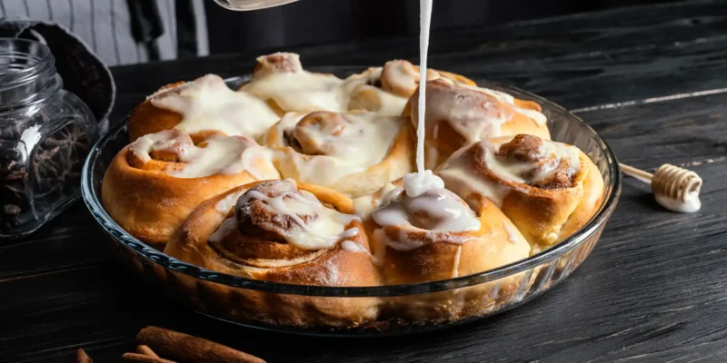 Ein hochauflösendes, dunkles Stimmungsbild zeigt eine Glasform, die mit frisch gebackenen, glasierten Cinnamon Rolls gefüllt ist. Eine Hand gießt aus einem durchsichtigen Gefäß eine dicke, weiße Glasur über die zimtigen Teigschnecken, wodurch der Guss auf einigen bereits glasierten Rolls glänzt. Auf dem rustikalen, dunklen Holztisch liegen Zimtstangen, ein Holzlöffel mit Honig und ein kleines Glasgefäß mit Kaffeebohnen, die das aromatische Thema unterstreichen.