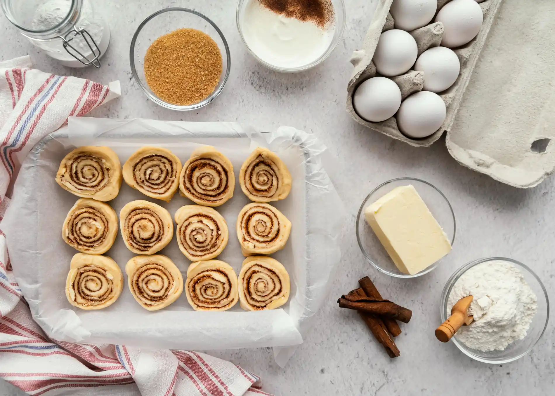 Ein von oben aufgenommenes Flat-Lay-Bild zeigt eine Backform mit 12 ungebackenen Cinnamon Rolls, die auf Backpapier in zwei Reihen angeordnet sind. Rund um die Form sind die Zutaten für das Gebäck ausgebreitet: Eine Schale mit braunem Zucker, eine mit weißem Mehl und einem Holzschaufel, eine mit Butter, eine Schale mit einer weißen Creme und Zimtpulver, eine halb volle Eierschachtel, ein Glasbehälter mit Mehl und Zimtstangen. Der gesamte Aufbau liegt auf einer hellen, strukturierten Oberfläche, die an Beton erinnert, und einer rot-weiß gestreiften Stoffserviette.